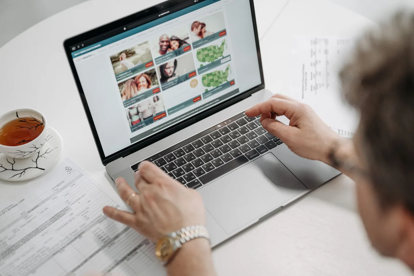 A person browsing a website on a laptop beside a cup of tea and papers.